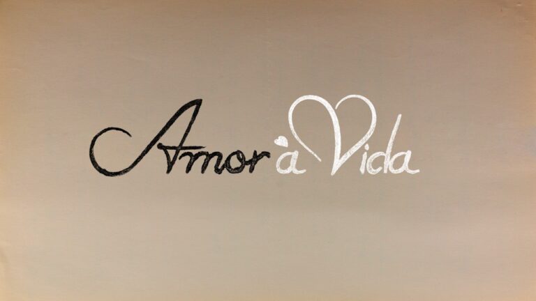 Logotipo da novela Amor à Vida com tipografia cursiva, palavra Amor em preto e Vida em branco com coração no topo da letra V, fundo bege claro clara