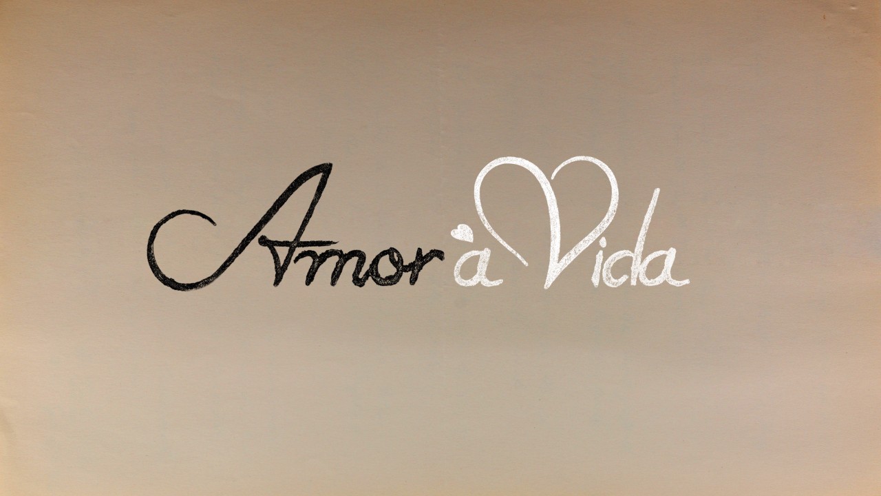Logotipo da novela Amor à Vida com tipografia cursiva, palavra Amor em preto e Vida em branco com coração no topo da letra V, fundo bege claro clara