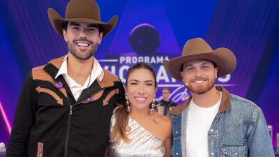 Luan Pereira, Patricia Abravanel e Gustavo Tubarão posam sorrindo no palco do Programa Silvio Santos com iluminação colorida ao fundo