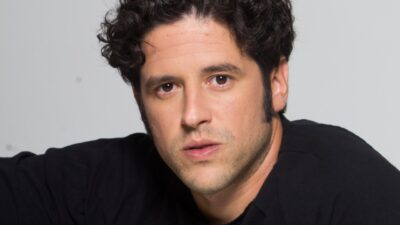 Ator Lucas Queiroga, conhecido como Lukete, possui cabelos pretos curtos e cacheados, olhos castanhos e veste camiseta preta em fundo cinza claro