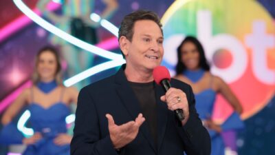 Luis Ricardo fala ao microfone no palco do Viva a Noite no SBT com dançarinas ao fundo e cenário colorido iluminado