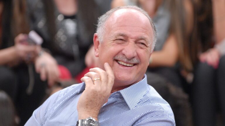 Luiz Felipe Scolari, o Felipão, sorri usando camisa social listrada de azul e branco, com a mão no queixo e relógio no pulso, no palco do Altas Horas