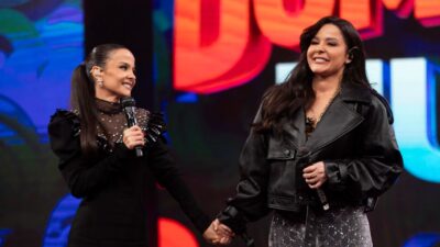 Cantoras Maiara e Maraisa sorriem de mãos dadas no palco do Domingão com Huck, segurando microfones e vestindo roupas pretas com brilhos e couro