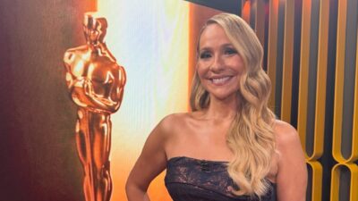 Maria Beltrão sorri na transmissão do Oscar 2026, usa vestido tomara que caia escuro, cabelos loiros ondulados, fundo com estatueta dourada gigante