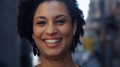 Marielle Franco sorri abertamente para a câmera em close, exibindo seus cabelos cacheados e brincos grandes e redondos, com fundo urbano desfocado