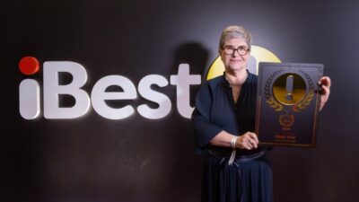 Marília Assef posa com placa de premiação iBest do Roda Viva diante de painel com logotipo iBest ao fundo