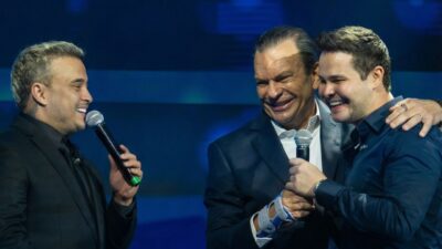 Matheus e Kauan sorriem ao lado de Roberto Sallouti em palco iluminado, segurando microfones e vestindo trajes sociais em tons de preto e azul escuro