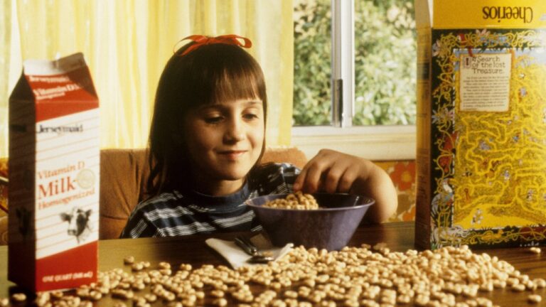 Matilda (Mara Wilson) pega cereal em tigela sobre mesa com leite e caixa de cereal em cena do filme Matilda