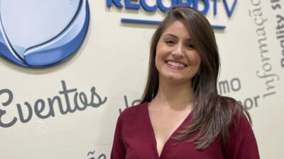 Natália Satler sorri com vestido vinho em frente a parede com logotipo da Record e palavras relacionadas a eventos