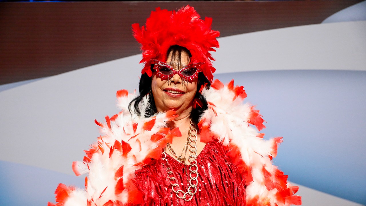Neísa Pantera sorri no programa The Noite, usa figurino vermelho com franjas, adereço de penas na cabeça, óculos de sol com brilhos e estola branca