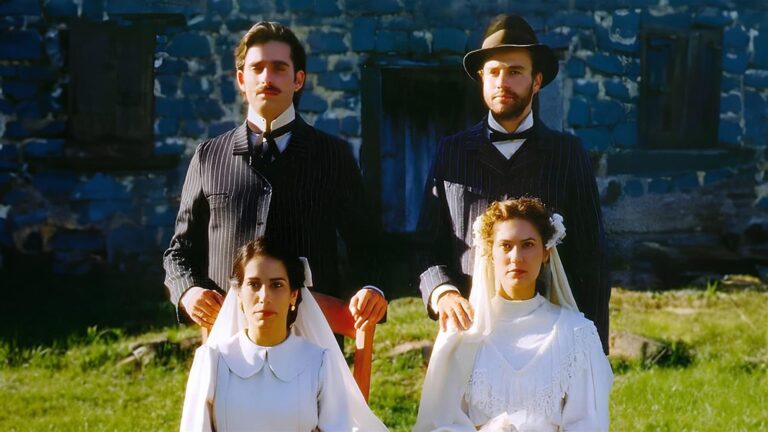 Dois casais vestidos com roupas de época, os homens usam ternos escuros e as mulheres vestidos de noiva brancos com véus, em frente a uma casa azul