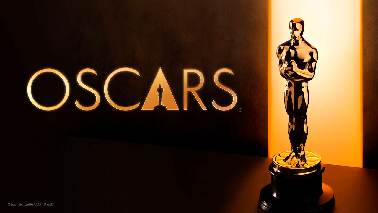 Estatueta dourada do Oscar posicionada à direita sobre base preta, logomarca Oscars iluminada em dourado ao centro, fundo escuro com feixe de luz lateral