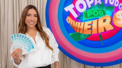 Patricia Abravanel sorri segurando várias notas de cem reais em leque, vestindo camisa branca diante do logo colorido do programa Topa Tudo por Dinheiro