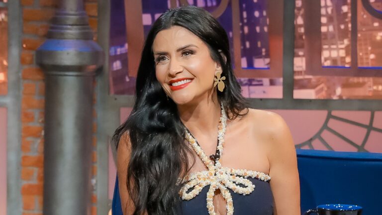 Patricia Salvador sorri no palco do The Noite, apresentadora usa vestido azul marinho com decote em laço de pérolas, brincos dourados e batom vermelho