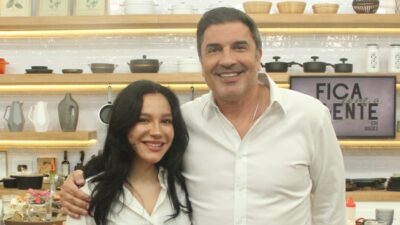 Priscilla e Edu Guedes sorriem abraçados no estúdio do Fica Com a Gente com bancada de cozinha e utensílios ao fundo