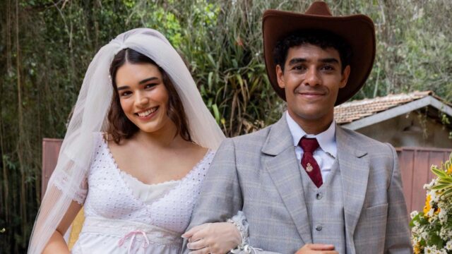Beatriz Anjinho como Pureza e Igor Fernandez como Picolé sorriem vestidos de noivos, ela usa véu e vestido branco rendado, ele traja terno cinza e chapéu marrom