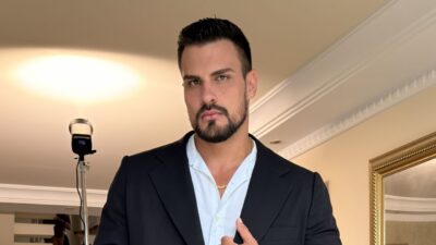 Renê Thristan em plano médio, veste blazer preto sobre camisa branca aberta e cordão de ouro, posando com olhar sério em ambiente interno iluminado