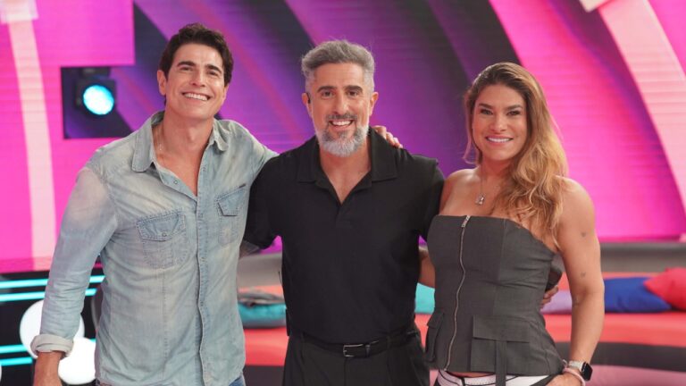 Reynaldo Gianecchini, Marcos Mion e Priscila Fantin sorrindo abraçados no palco do Caldeirão com cenário colorido ao fundo