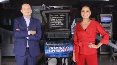 Roberto Cabrini veste terno azul ao lado de Carolina Ferraz, que usa conjunto vermelho, ambos sorriem em frente a um teleprompter da Record