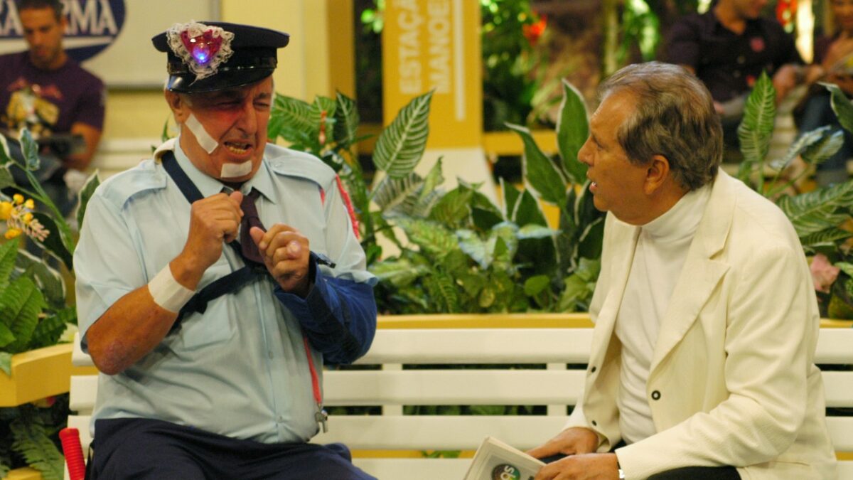 Roberto Marquis caracterizado de policial com curativos conversa com Carlos Alberto de Nóbrega em banco de A Praça É Nossa
