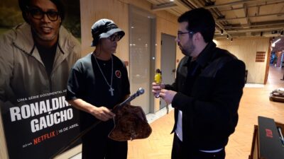 Ronaldinho Gaúcho com chapéu e colar segura bastão e bolsa enquanto conversa com Chico Pedrotti em evento da Netflix sobre série documental