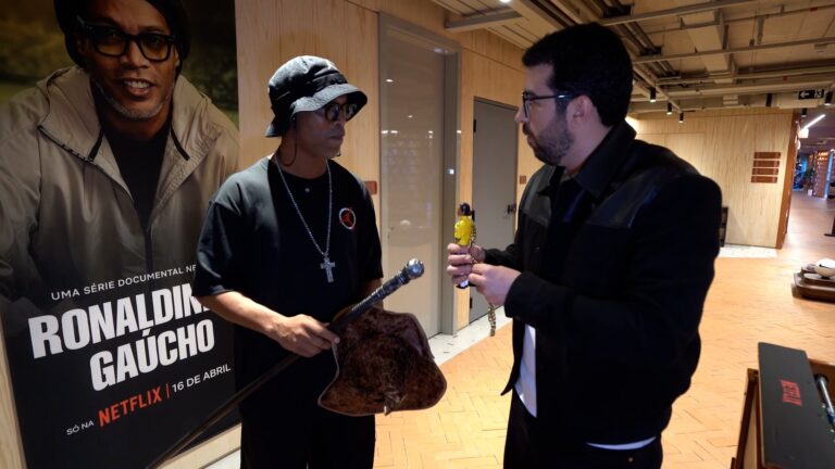 Ronaldinho Gaúcho com chapéu e colar segura bastão e bolsa enquanto conversa com Chico Pedrotti em evento da Netflix sobre série documental