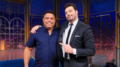 Ronaldo Fenômeno de camisa azul e Danilo Gentili de terno e gravata sorriem abraçados no cenário do programa The Noite, do SBT, com janelas ao fundo