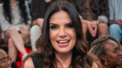 Jornalista Sabina Simonato sorri com cabelos castanhos ondulados e soltos, veste blusa preta e aparece em primeiro plano com a plateia ao fundo no Altas Horas