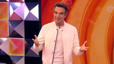 Tadeu Schmidt sorri com braços abertos, veste jaqueta bomber branca sobre camiseta clara e gesticula no cenário colorido e geométrico do reality BBB 26