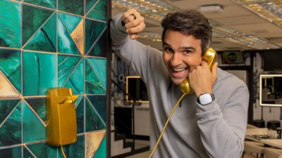 Apresentador Tadeu Schmidt sorri e segura o telefone dourado do BBB 26, veste blusa cinza de manga longa e apoia o braço em parede verde