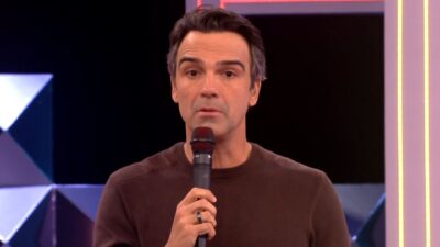 Tadeu Schmidt segura microfone preto, veste blusa marrom e encara a câmera com expressão séria no cenário iluminado do reality show BBB 26