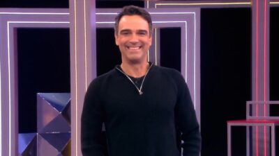 Tadeu Schmidt sorri no palco do BBB 26, veste blusa preta de gola alta com zíper, cenário ao fundo possui luzes e estruturas neon