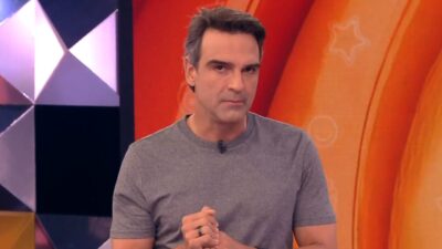 Tadeu Schmidt veste camiseta cinza, mãos juntas, no cenário do BBB 26, com fundo laranja vibrante e elementos geométricos coloridos