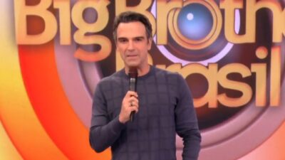 Tadeu Schmidt de blusa azul quadriculada segura microfone e fala no palco do BBB 26, com o logotipo do reality show da Globo ao fundo