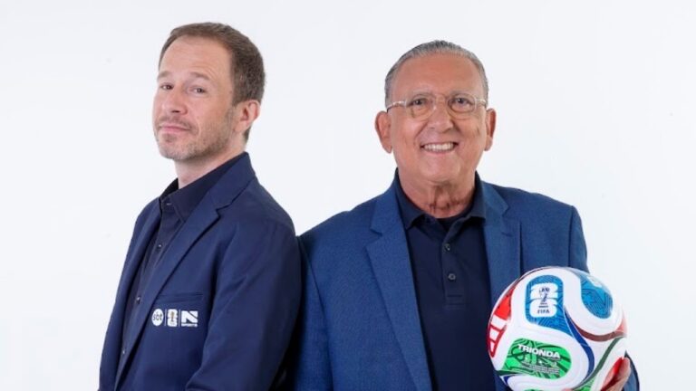 Tiago Leifert e Galvão Bueno posam juntos, Galvão segura bola de futebol da Copa do Mundo, ambos vestem paletós azuis e camisas escuras, fundo branco neutro