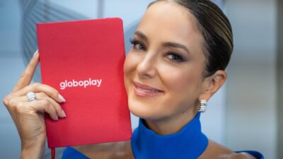 Apresentadora Ticiane Pinheiro sorri segurando um caderno vermelho com o logotipo do Globoplay, veste blusa azul vibrante e usa brincos prateados