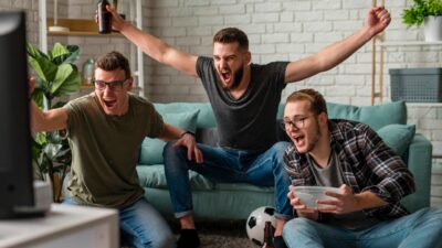 Torcedores comemoram gol assistindo jogo de futebol na TV em sala com sofá bola no chão e garrafa de bebida nas mãos