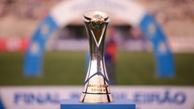Troféu do Brasileirão Série D em prata e dourado posicionado no centro do gramado, com a inscrição da competição na base e um arco azul ao fundo