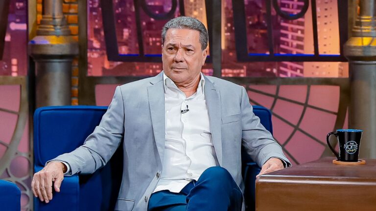 Vanderlei Luxemburgo sentado em poltrona azul no programa The Noite, veste paletó cinza e camisa branca, com caneca preta da atração sobre mesa de madeira