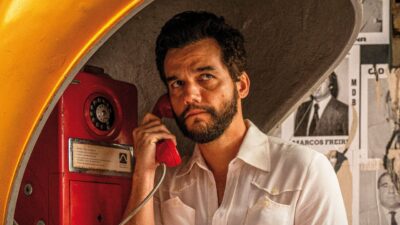 Wagner Moura de barba e camisa branca atende telefone público vermelho dentro de um orelhão amarelo, com cartazes políticos ao fundo em O Agente Secreto