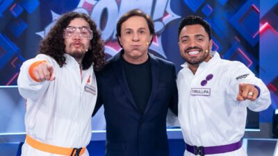 Whindersson Nunes e Tirullipa vestem macacões brancos e apontam para frente, enquanto Tom Cavalcante faz bueiro de terno azul no cenário do game show Boom!