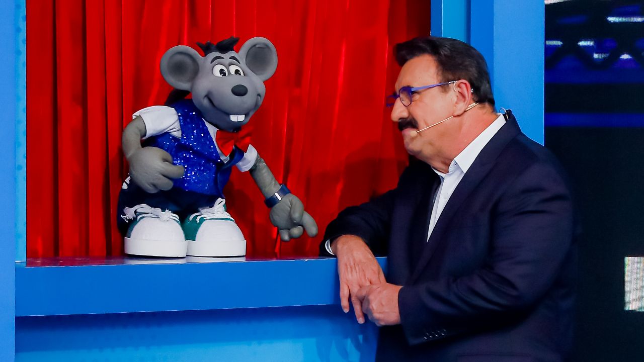 Boneco Xaropinho veste colete azul e gravata borboleta vermelha ao lado do apresentador Ratinho, que traja terno escuro, no cenário do Programa do Ratinho
