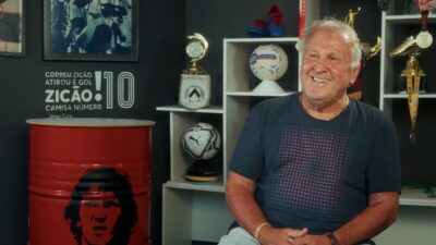 Zico sorri sentado em estúdio com prateleiras de troféus e bolas ao fundo, ao lado de barril vermelho decorado com seu rosto e a frase Zicão Camisa 10