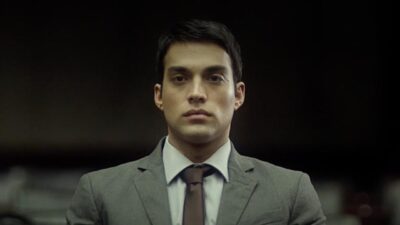 Alex interpretado por James Floyd encara à frente com expressão séria usando terno e gravata em cena o filme O Novato