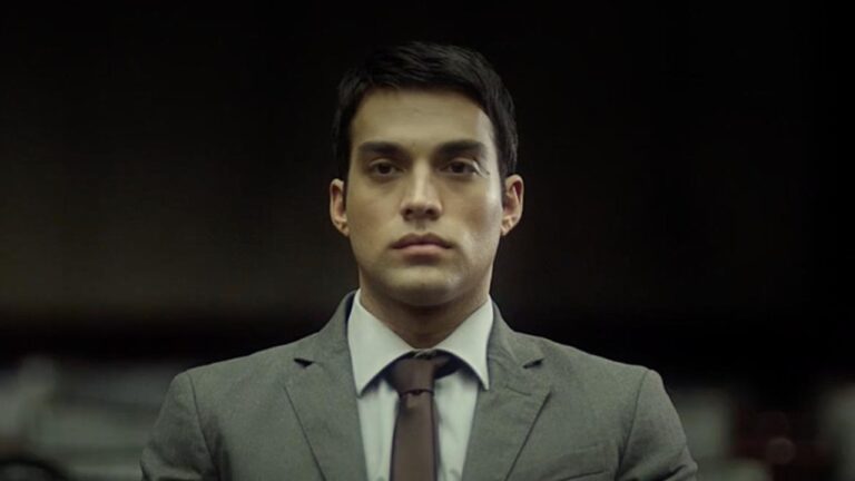 Alex interpretado por James Floyd encara à frente com expressão séria usando terno e gravata em cena o filme O Novato