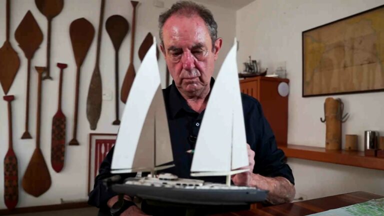 Amir Klink segura maquete de veleiro sobre mesa em ambiente interno com remos decorativos na parede ao fundo