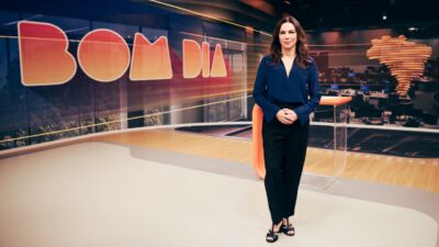 Ana Paula Araújo posa no estúdio do Bom Dia Brasil com telão ao fundo exibindo o nome do telejornal e redação ao fundo