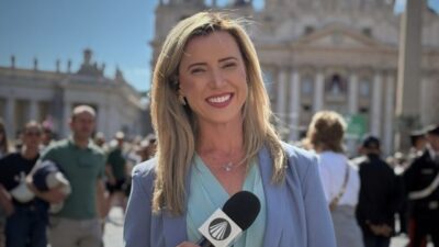 Camila Morais segura microfone da TV Aparecida durante reportagem em praça com igreja histórica e fiéis ao fundo