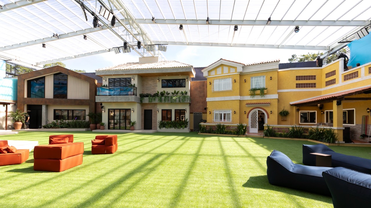 Área externa do Casa do Patrão com gramado amplo, sofás espalhados e fachadas das casas ao fundo sob cobertura de estúdio