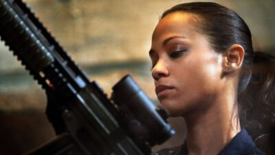 Cataleya interpretada por Zoe Saldaña segura rifle com mira e observa arma em cena do filme Colombiana: Em Busca de Vingança